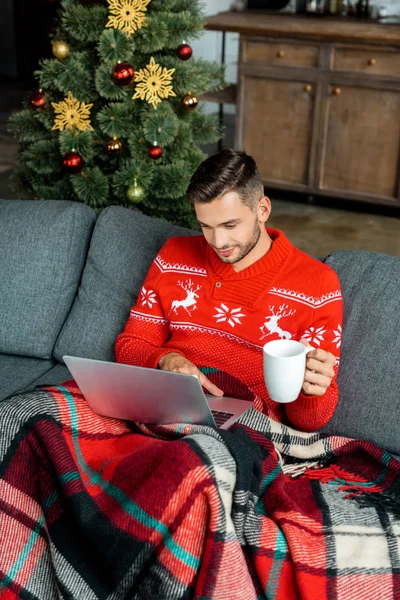 Hombre joven con taza de café sentado en el sofá debajo de la manta y el uso de la computadora portátil cerca del árbol de Navidad en casa - foto de stock