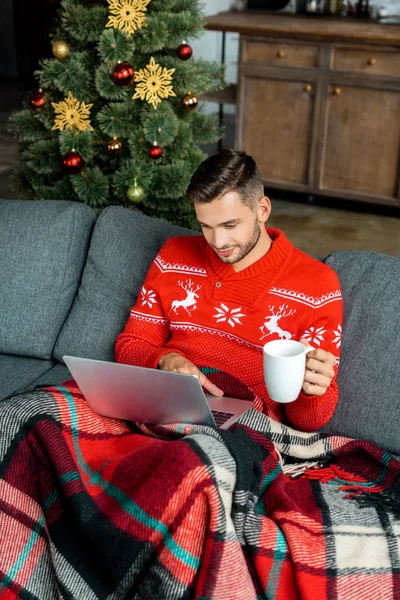 Giovane uomo con tazza di caffè seduto sul divano sotto coperta e utilizzando il computer portatile vicino all'albero di Natale a casa — Foto stock