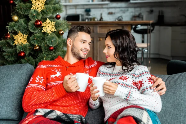 Coppia sorridente con tazze di caffè seduti sul divano sotto coperta vicino all'albero di Natale a casa — Foto stock