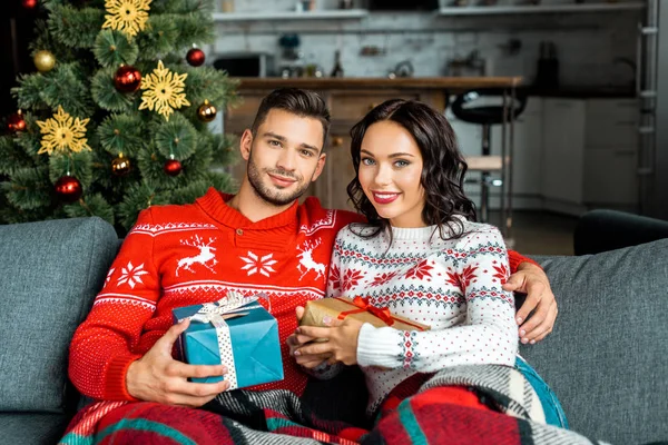 Alegre pareja sentada en el sofá con cajas de regalo cerca del árbol de Navidad en casa - foto de stock