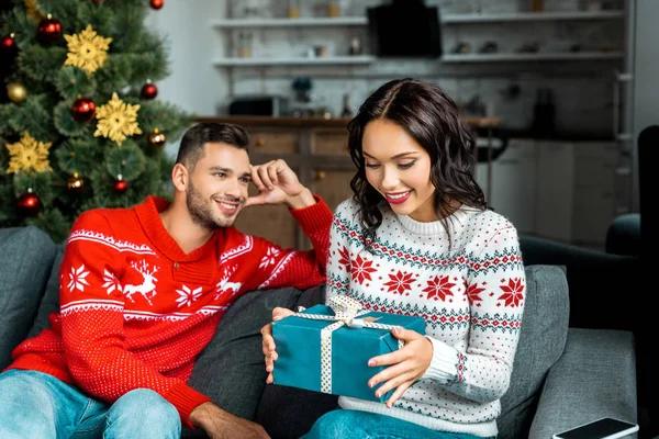Attractive young woman untying christmas present while her boyfriend sitting on sofa behind at home — Stock Photo
