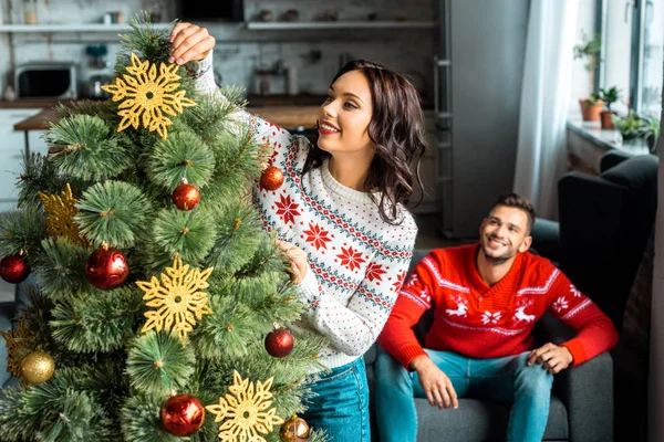 Selektiver Fokus der Frau, die den Weihnachtsbaum schmückt, während ihr Freund zu Hause auf dem Sofa sitzt — Stockfoto