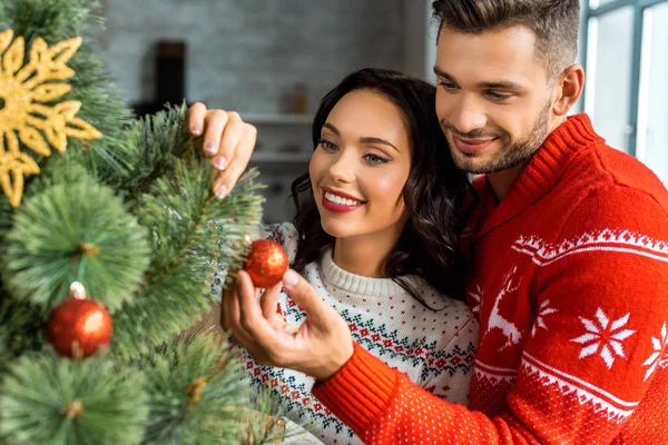 Smiling young couple decorating christmas tree by baubles at home — Stock Photo