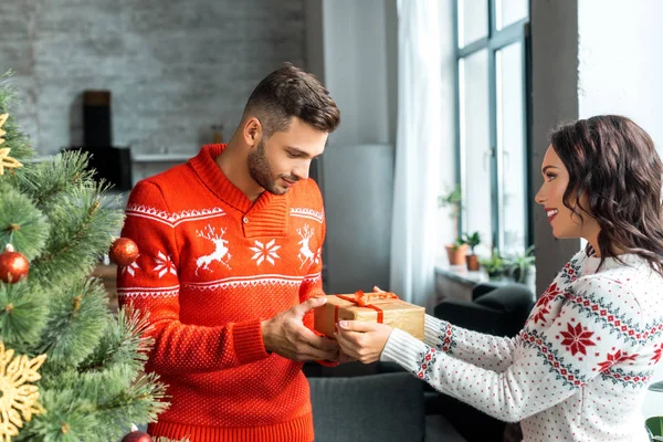 Vue latérale de la boîte cadeau femme heureuse au petit ami près de l'arbre de Noël à la maison — Photo de stock