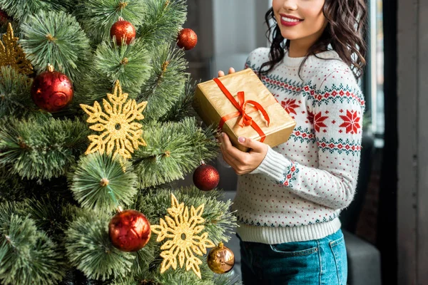 Partial view of young woman holding gift box near christmas tree at home — Stock Photo