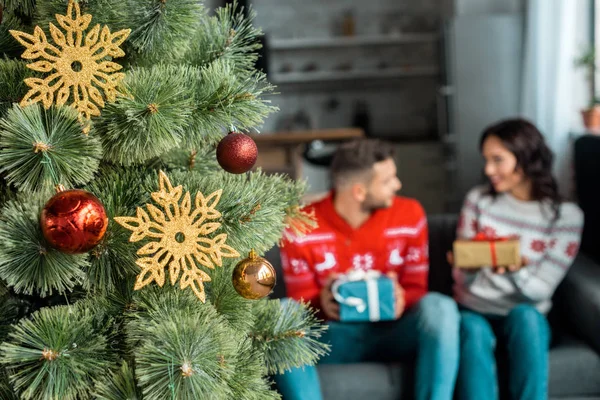 Selective focus of decorated christmas tree and couple with gift boxes sitting on sofa behind at home — Stock Photo