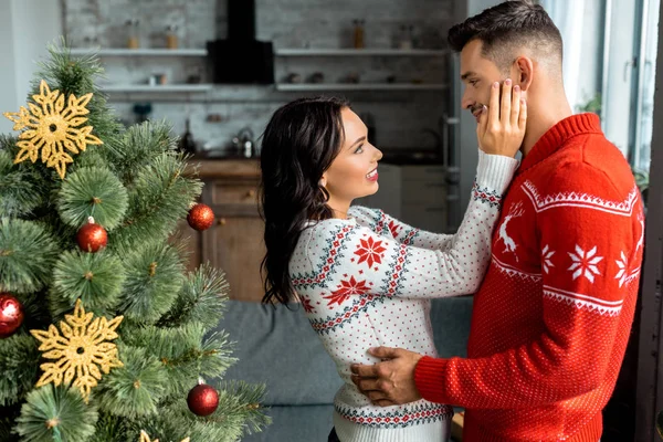 Seitenansicht eines Paares, das sich in der Nähe des Weihnachtsbaums zu Hause umarmt und ansieht — Stockfoto