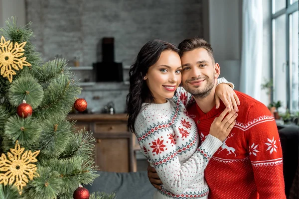 Retrato de jovem casal abraçando e olhando para a câmera perto da árvore de natal em casa — Fotografia de Stock