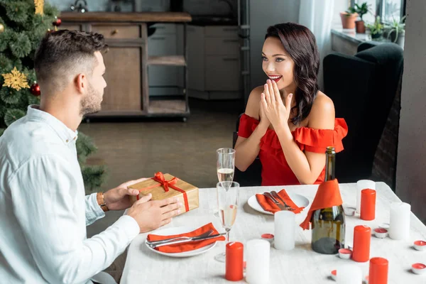 Side view of man gifting christmas present box to excited girlfriend sitting at served table at home — Stock Photo