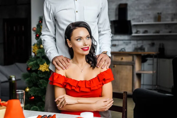 Image recadrée de l'homme embrassant petite amie épaules alors qu'elle est assise à la table de Noël servi et regardant loin à la maison — Photo de stock