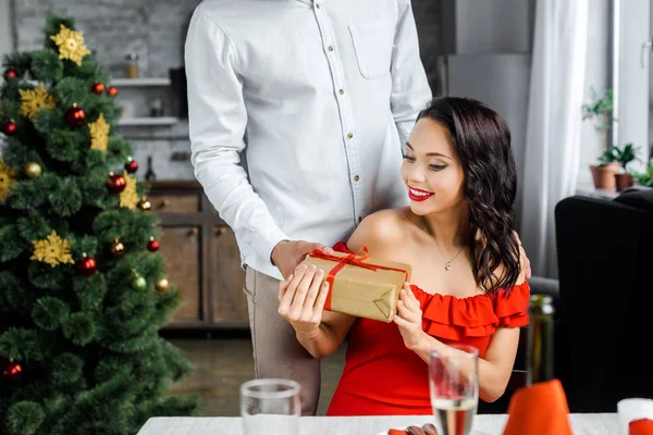 Vista parziale dell'uomo regalando scatola regalo alla fidanzata sorridente seduta al tavolo servito a casa — Foto stock