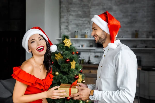 Uomo sorridente in cappello di Natale regalo regalo scatola a ridere fidanzata a casa — Foto stock