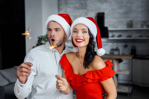 Emotional happy couple in christmas hats celebrating with sparkles at home — Stock Photo