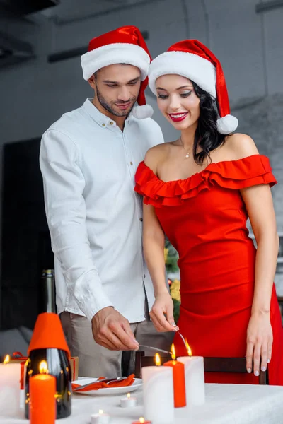 Jeune couple dans les chapeaux de Noël brûlant bâtons d'éclat par des bougies à la maison — Photo de stock