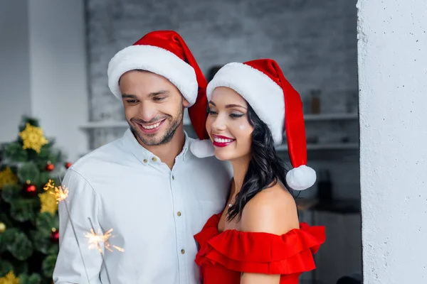 Jovem casal alegre em chapéus de Natal comemorando com brilhos em casa — Fotografia de Stock