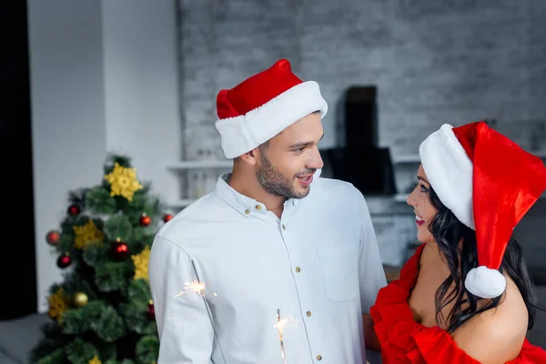 Smiling young couple in christmas hats holding bengal lights and looking at each other at home — Stock Photo