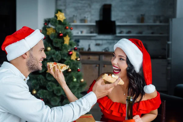 Laughing couple in chrtistmas hats feeding each other by pizza at home — Stock Photo