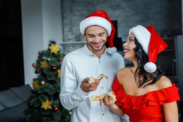 Enfoque selectivo de la pareja joven en sombreros de Navidad comer pizza en casa - foto de stock