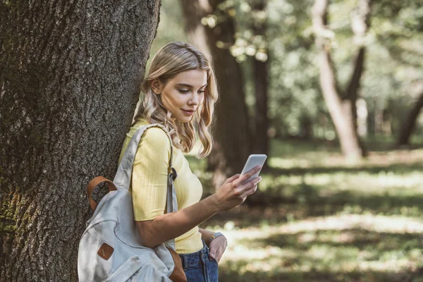 Mujer joven con mochila en el hombro usando teléfono inteligente en el bosque - foto de stock