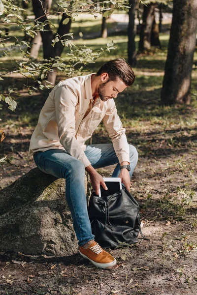 Joven poniendo tableta digital en la mochila en el parque - foto de stock