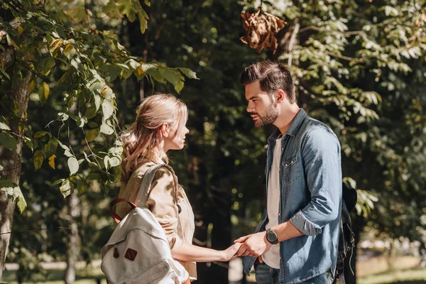 Вид сбоку молодой пары с рюкзаками, держащейся за руки и смотрящей друг на друга в парке — стоковое фото