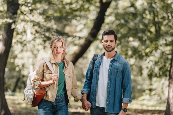 Portrait of young couple with backpacks holding hands and looking at camera in park — Stock Photo