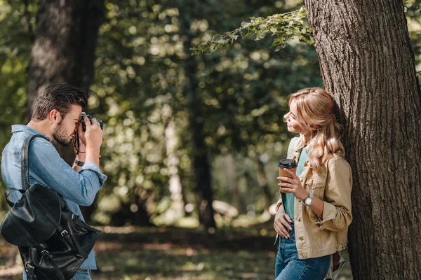 Side view of man taking picture of smiling girlfriend with coffee to go in park — Stock Photo