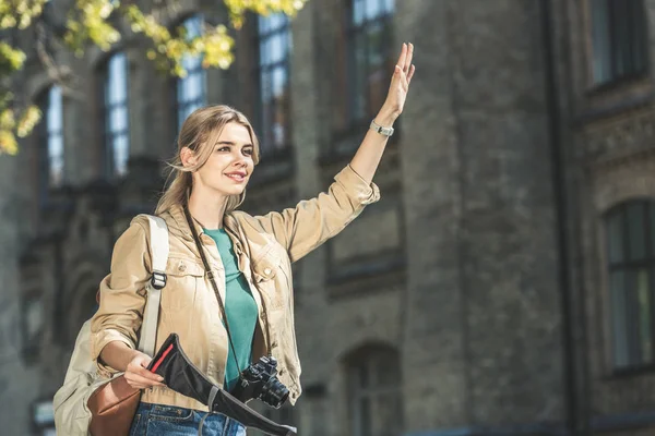 Portrait de jeune voyageur avec sac à dos, appareil photo et carte appelant taxi dans la rue — Photo de stock