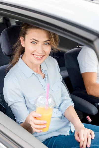 Hermosa mujer feliz con taza de plástico de jugo de naranja sentado en el coche - foto de stock