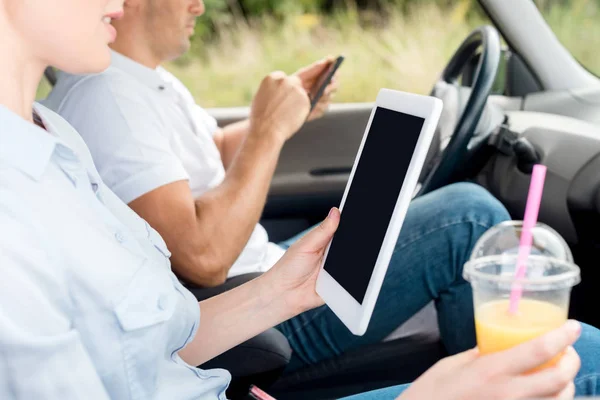 Cropped shot of woman with plastic cup of orange juice using tablet in car while man using smartphone on background — Stock Photo