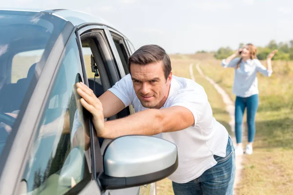 Man pushing broken car while his wife talking by phone in field during car trip — Stock Photo
