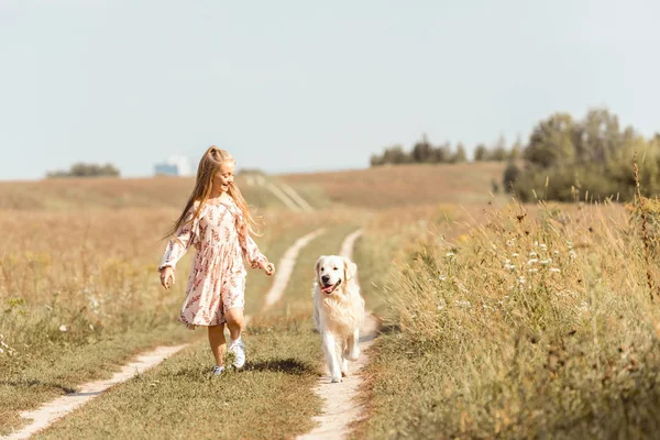Beautiful little child running with adorable golden retriever dog in field — Stock Photo