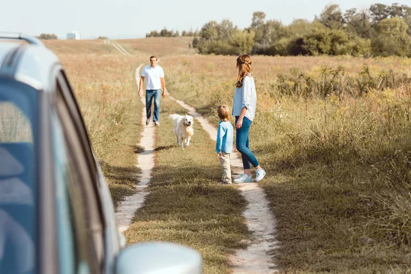 Hermosa familia con perro caminando por la carretera de campo junto con el coche en primer plano - foto de stock