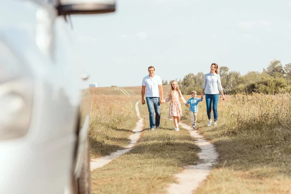 Belle jeune famille tenant la main et marchant à la voiture par champ — Photo de stock