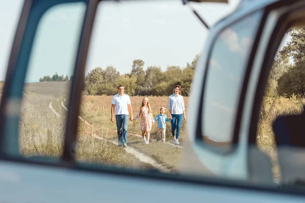 Счастливая молодая семья, держащаяся за руки и идущая к машине по полю — стоковое фото