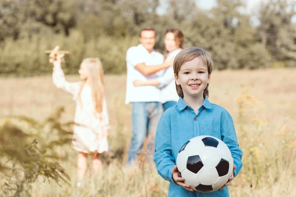 Чарівна маленька дитина тримає м'яч, а його сім'я стоїть розмитим на фоні в полі — стокове фото