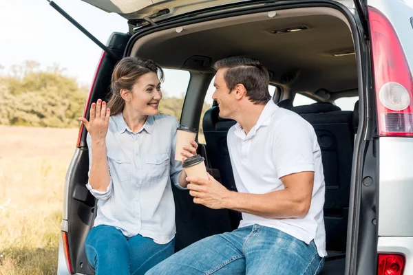 Happy adult couple with paper cups of coffee sitting in car trunk while travelling by car — Stock Photo