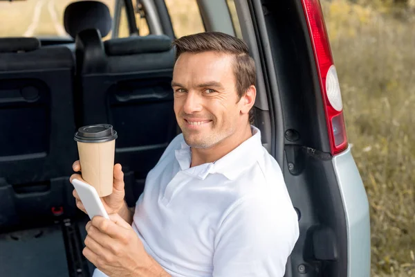 Hombre feliz con taza de papel de café sentado en el maletero del coche y mirando a la cámara mientras se utiliza el teléfono inteligente - foto de stock