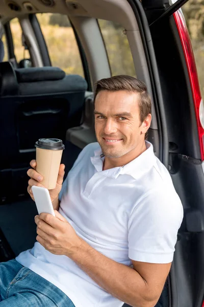 Улыбающийся человек с бумажной чашкой кофе сидит в багажнике автомобиля и смотрит в камеру во время использования смартфона — стоковое фото
