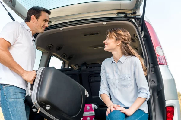 Happy adult man packing luggage into car trunk while his girlfriend looking at him — Stock Photo