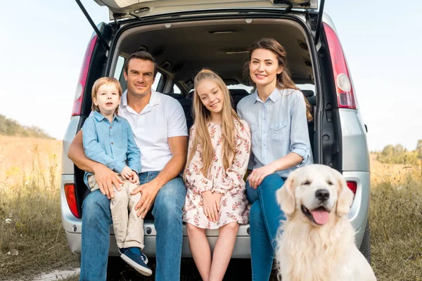 Hermosa familia joven con perro recuperador sentado en el maletero del coche y mirando a la cámara en el campo - foto de stock