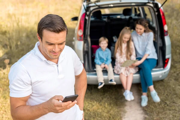 Homme heureux en utilisant smartphone tandis que sa famille assis dans le coffre de la voiture dans le domaine — Photo de stock