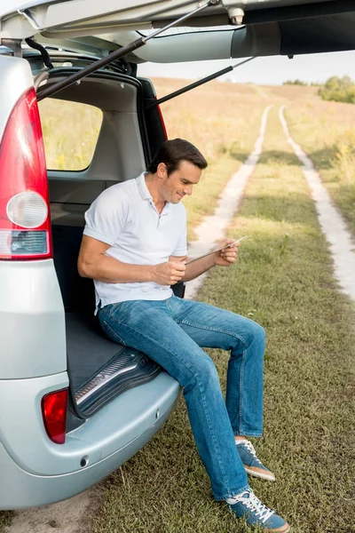 Bell'uomo sorridente seduto nel bagagliaio dell'auto e guardando la mappa in campo — Foto stock