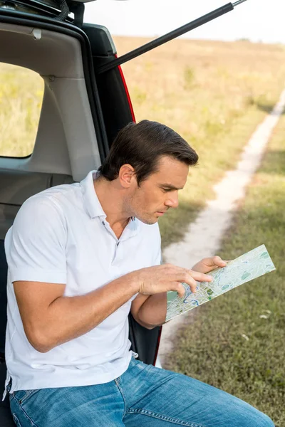 Homme attrayant assis dans le coffre de la voiture et naviguer avec carte dans le champ — Photo de stock