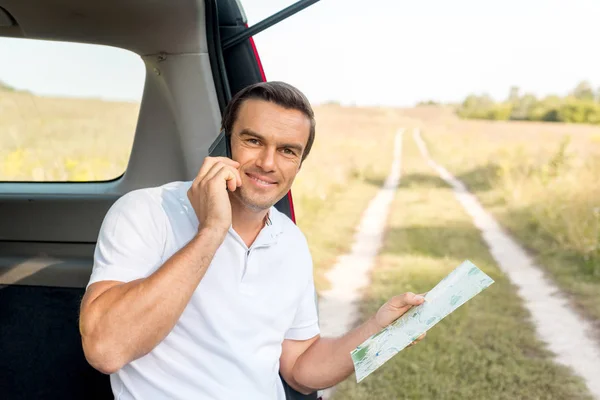 Handsome man sitting in car trunk with map and talking by phone in field — Stock Photo