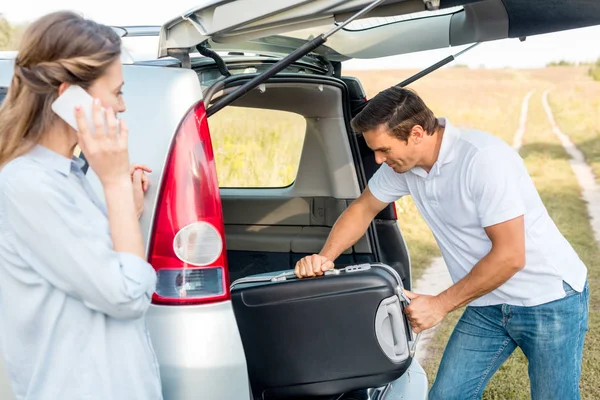 Hombre adulto feliz embalaje de equipaje en el maletero del coche mientras su novia habla por teléfono en el campo - foto de stock