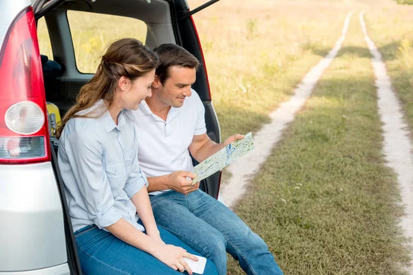 Sonriente pareja adulta sentada en el maletero del coche y mirando el mapa mientras tiene viaje en coche - foto de stock