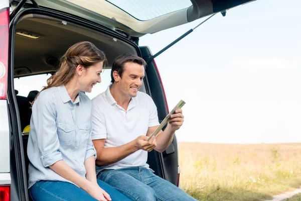 Heureux couple adulte assis dans le coffre de la voiture et en regardant la carte tout en ayant voyage en voiture — Photo de stock