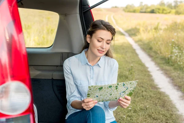 Beautiful adult woman sitting in car trunk and looking at map in field — Stock Photo