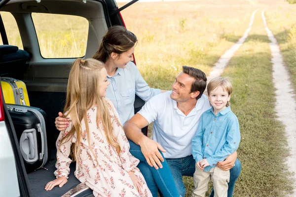 Hermosa familia joven sentada en el maletero del coche y abrazándose en el campo mientras tiene viaje en coche - foto de stock